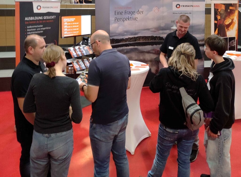 Ein Mitarbeiter von Franken Guss spricht am Messestand mit einem Mann und einer Jugendlichen. Ein anderer Mitarbeiter spricht mit einer Frau und einem Jugendlichen.